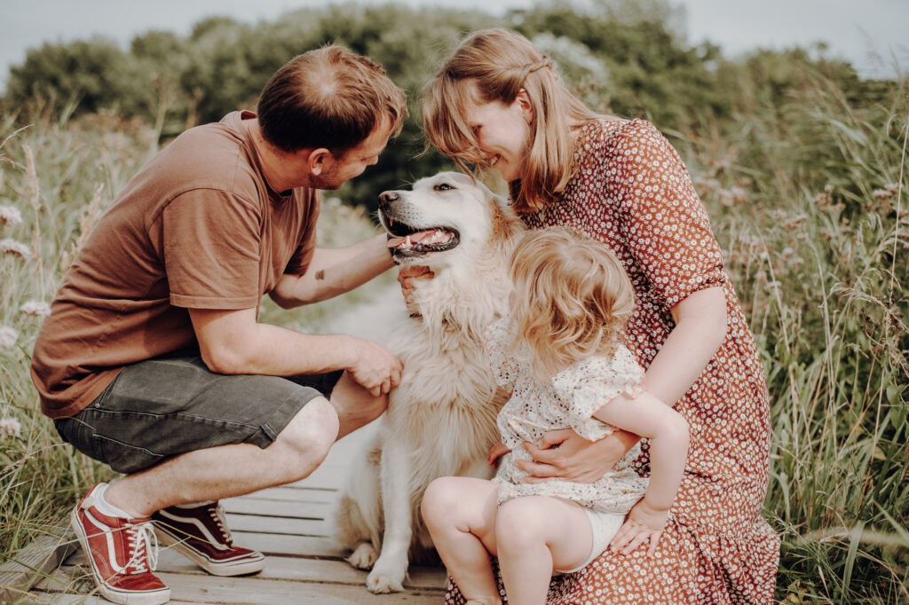 Familie mit kleinem Kind und einem Golden Retriever sitzt lächelnd auf einem Holzweg in der Natur.