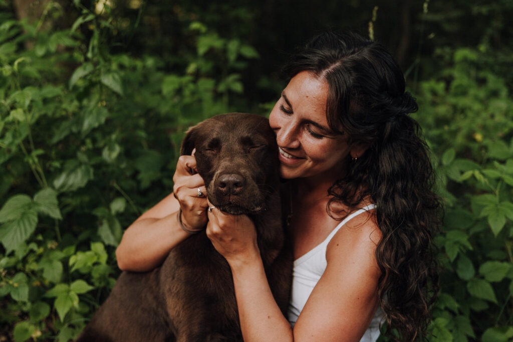 Frau schmiegt ihr Gesicht lächelnd an die Wange ihres braunen Labradors, der genießerisch die Augen schließt.